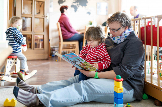Familienarbeit im Kirchenkreis Neustadt-Wunstorf. Ehrenamtliche ließt Kind ein Buch vor. Mutter im Hintergrund im Geschäftsgespräch.